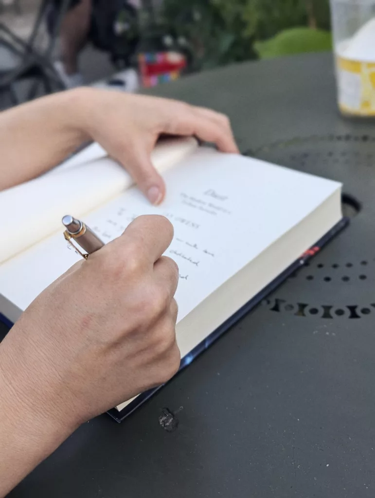 A pair of hands writing a message with a silver pen on the title page of a hardcover book on an outdoor table.
