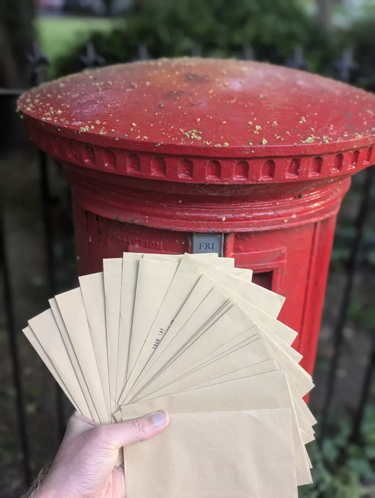 A hand holding a fanned-out stack of brown paper envelopes in front of a red British pillar box.