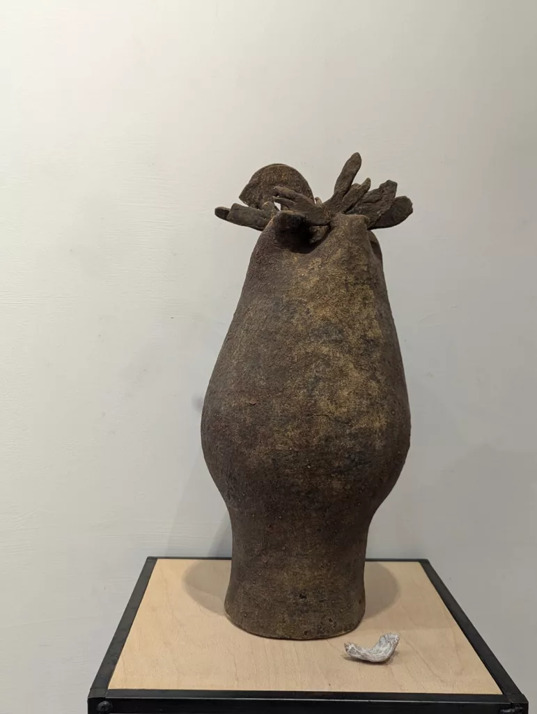 A dark, textured ceramic vessel with a crown of irregular protrusions, sitting on a light wood plinth against a white gallery wall.