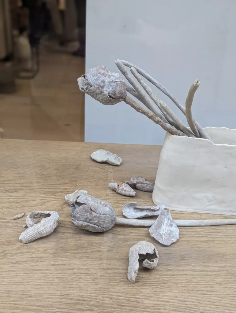 Textured grey ceramic tulips in a white handmade vase, with one flower and broken petals scattered across a light wooden table.