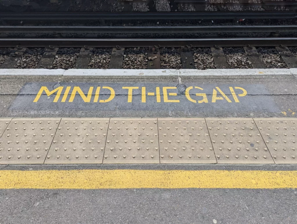 Yellow stenciled MIND THE GAP warning on a train platform edge above railway tracks, with tactile paving and a yellow safety line.