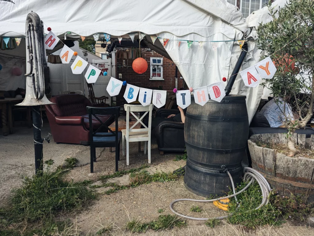 Happy Birthday banner and bunting across a marquee entrance featuring vintage chairs, a large water butt, and a tuba mounted to a post.