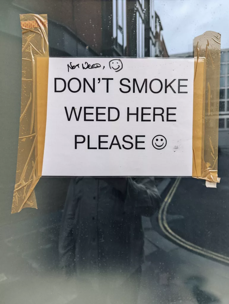 A sign taped to a window reads "DON'T SMOKE WEED HERE PLEASE :)" with a handwritten note above it saying "NOT WEED, :)