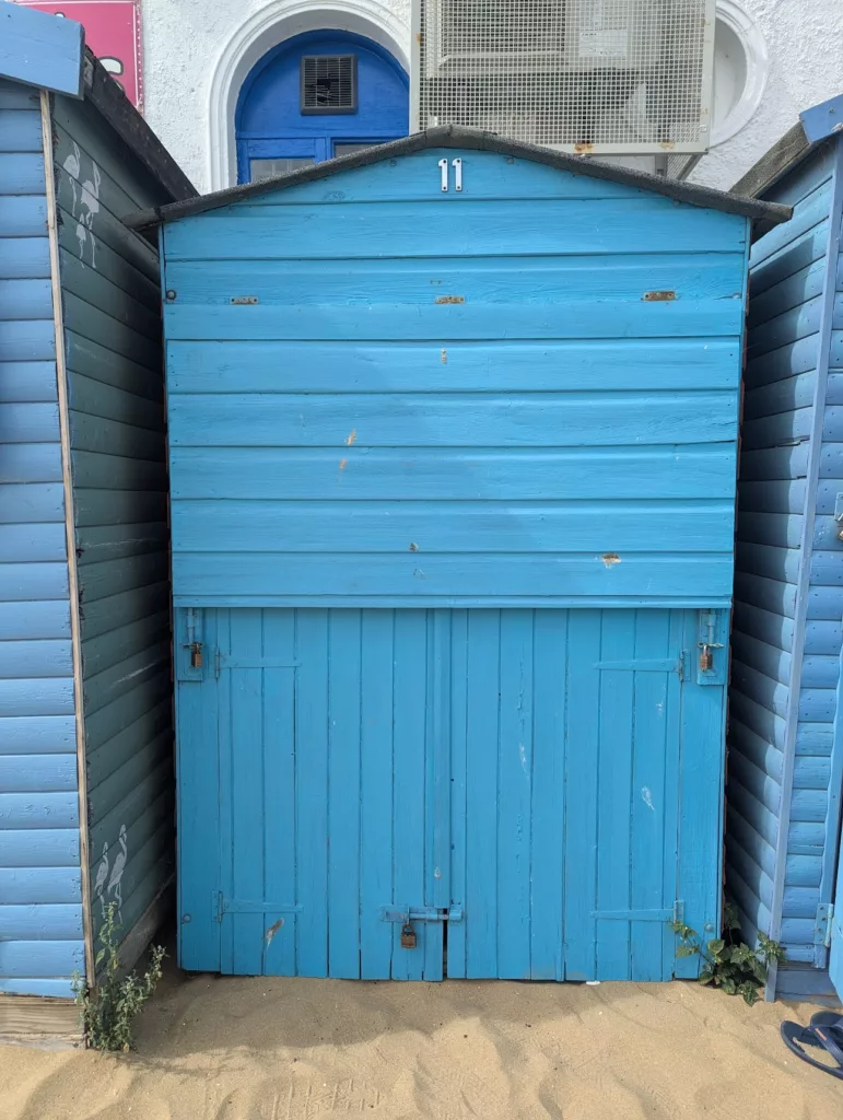A bright blue wooden beach hut numbered 11 on sand, positioned between two similar huts with a white building and blue arched window behind.