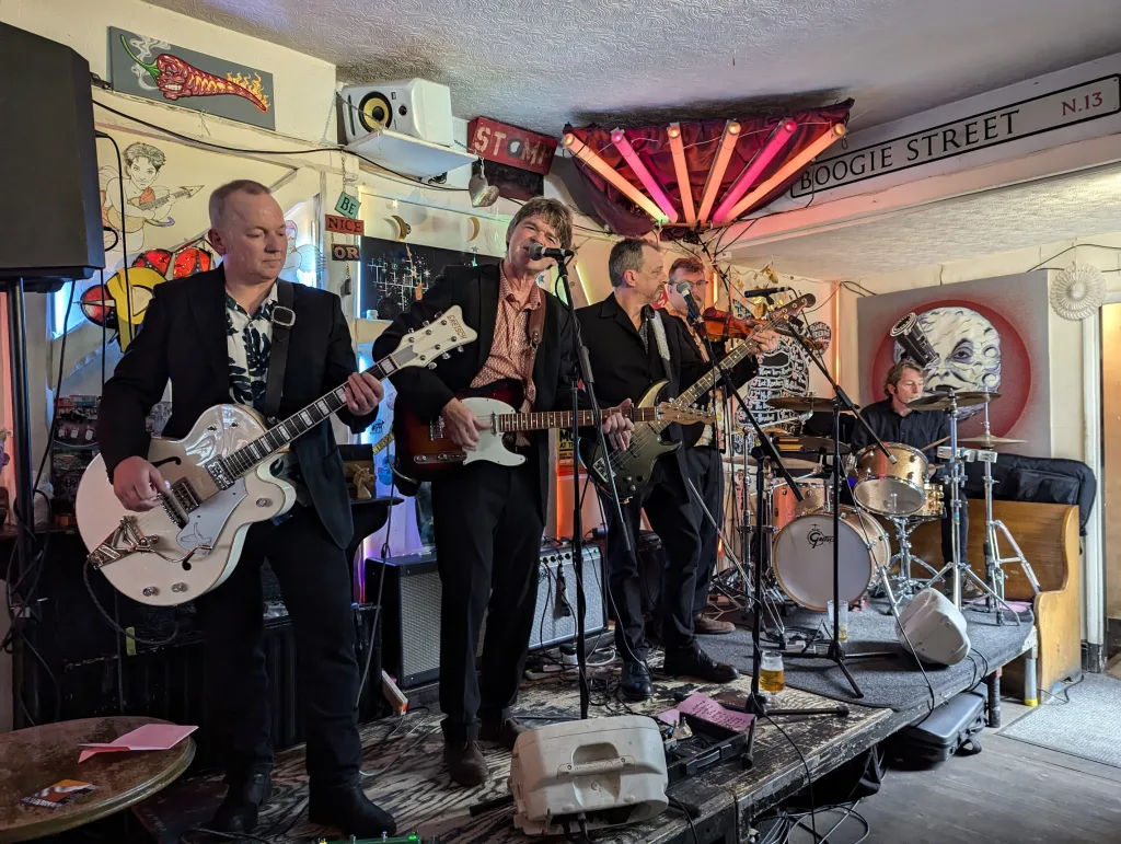 A five-piece band performing live on a small pub stage decorated with red neon lights and a sign reading Boogie Street.