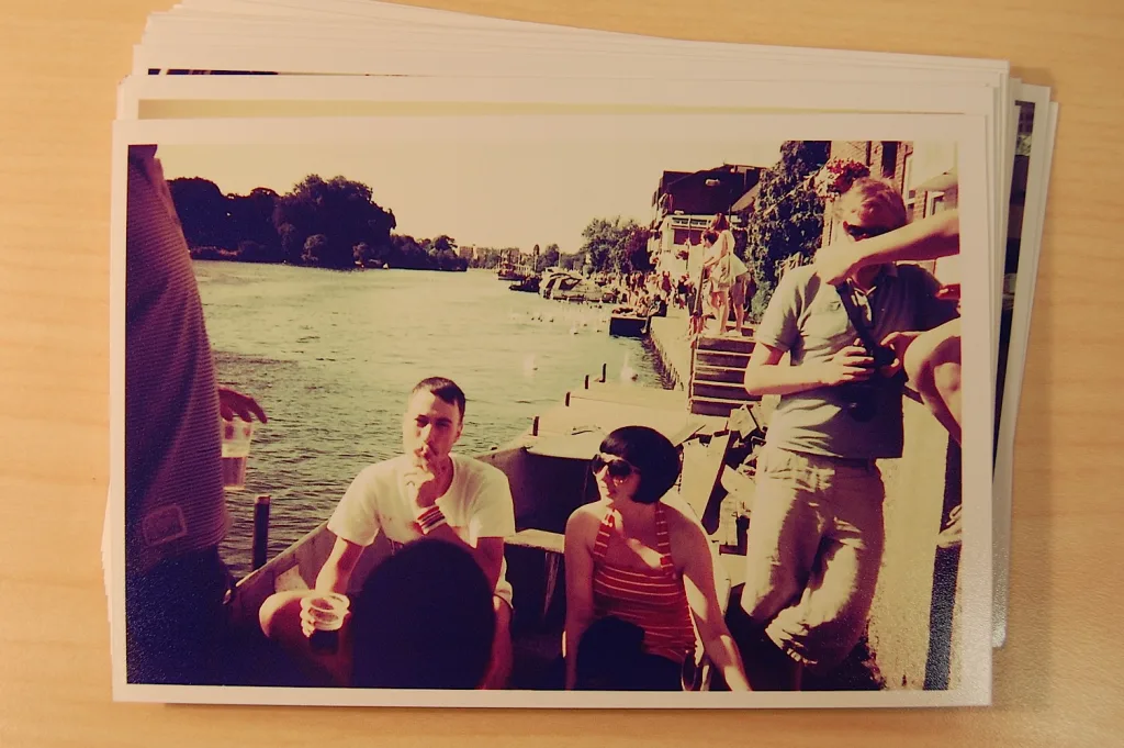 A stack of printed photos on a wooden surface. The top photo depicts friends relaxing by a riverbank in warm, vintage-toned sunlight.