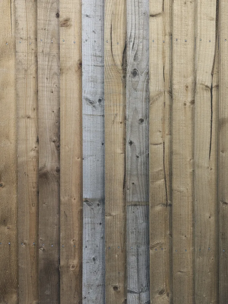 Vertical wooden fence planks in varying shades of tan and weathered grey, showing natural grain, knots, and small nail heads.