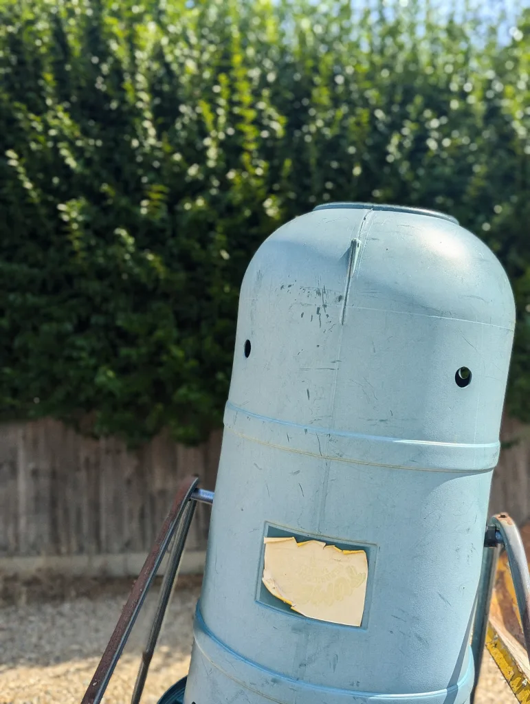 A light blue plastic garden bin with two small holes that look like eyes, standing outdoors in front of a green hedge.