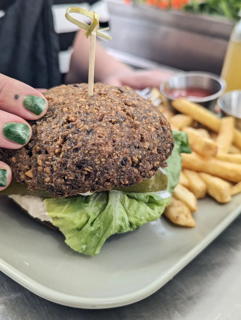 A hand with green glitter nails holds a dark seeded burger with lettuce and pickles, served with fries and ketchup on a pale plate.