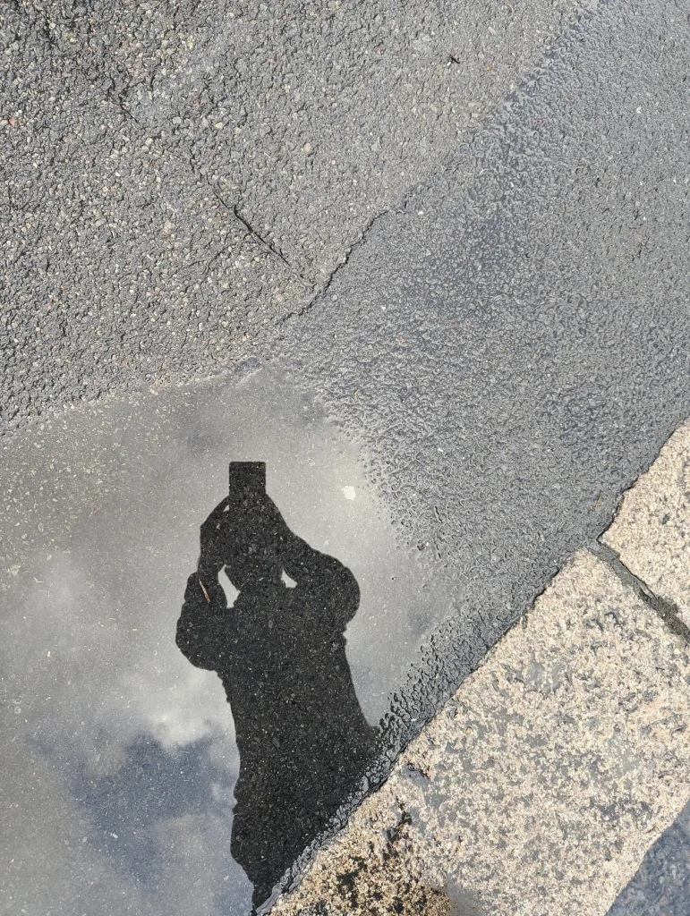 Reflection of a person holding a smartphone in a puddle on grey asphalt next to a textured stone curb under a cloudy sky.