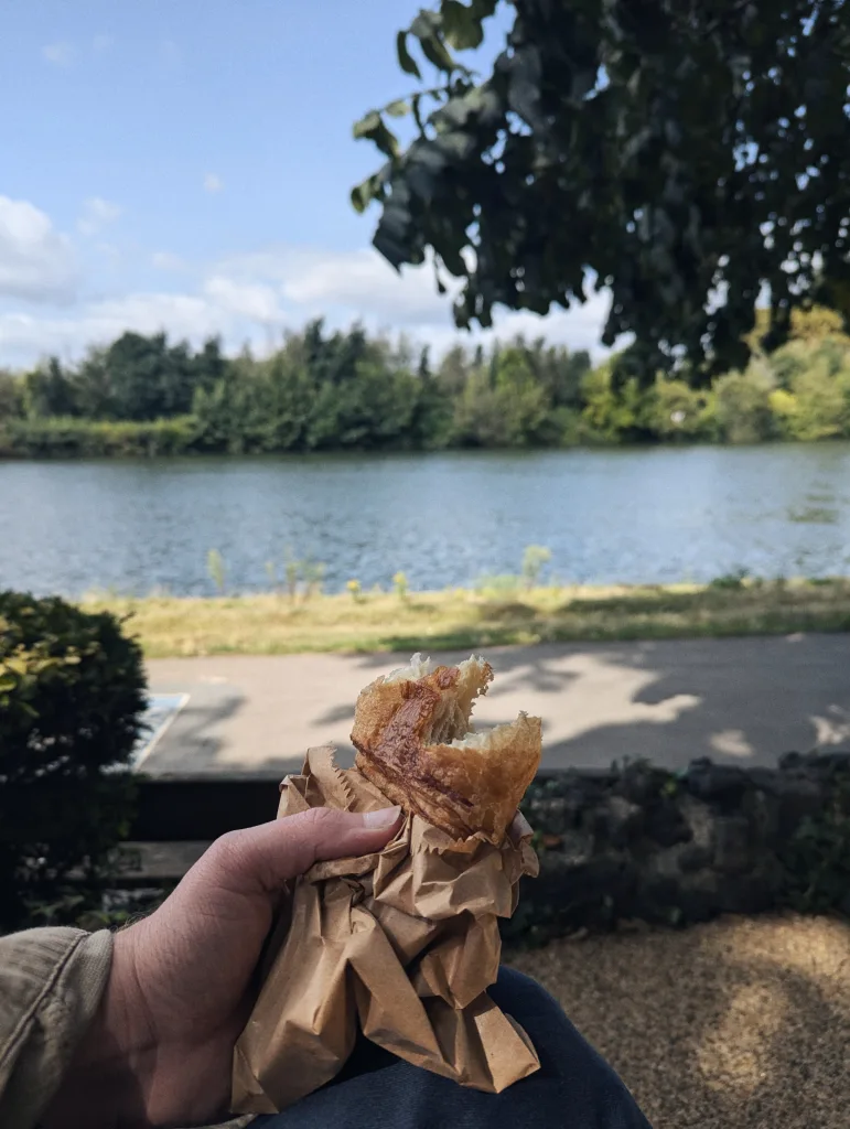 A hand holds a partially eaten flaky pastry in a brown paper bag overlooking a calm lake and wooded shoreline on a sunny day.