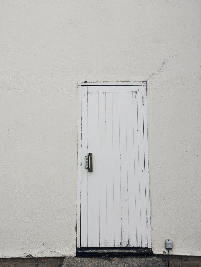 A weathered white wooden door with a digital keypad lock, set into a plain white rendered wall with a small crack.