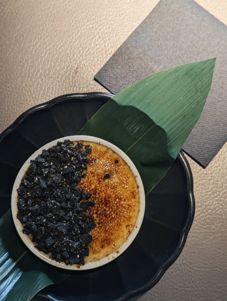 Crème brûlée with a black sesame crumble topping, served in a white ramekin on a black scalloped plate with a large green leaf.