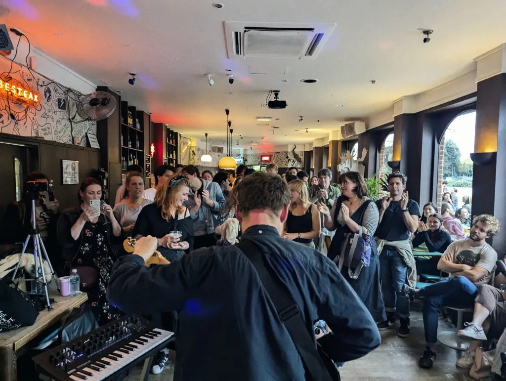 A musician seen from behind performs with a keyboard in a crowded bar, while an audience watches and applauds under ambient lighting.