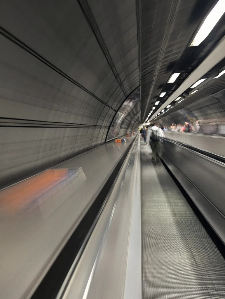 Motion-blurred long exposure of a person on a moving walkway in a grey, curved transit tunnel with bright ceiling lights.
