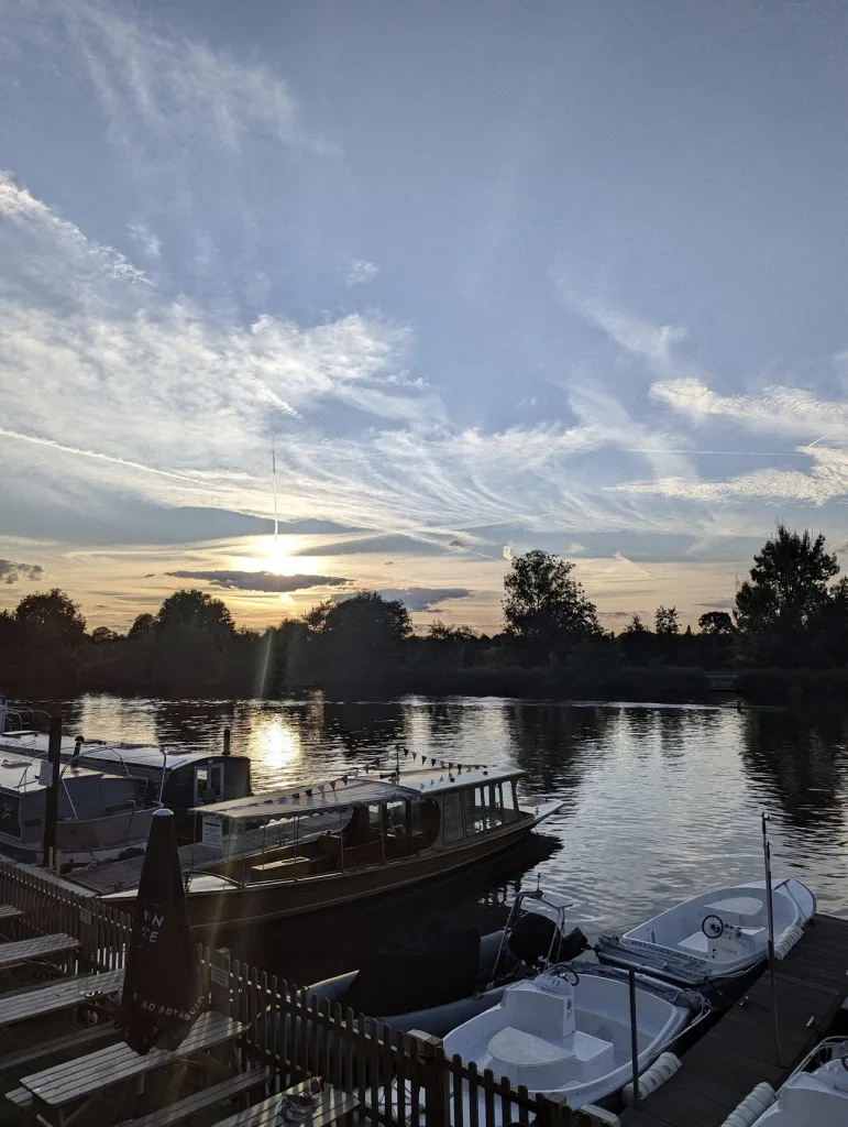 Sunset over a river with boats moored at a dock, including a wooden tour boat with bunting, under a sky of wispy clouds.