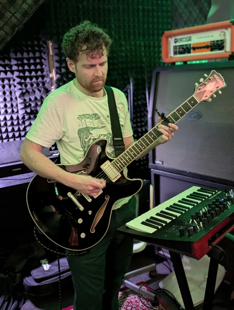 A man playing a semi-hollow electric guitar in a music studio with green lighting, an Orange amplifier, and a synthesizer keyboard.