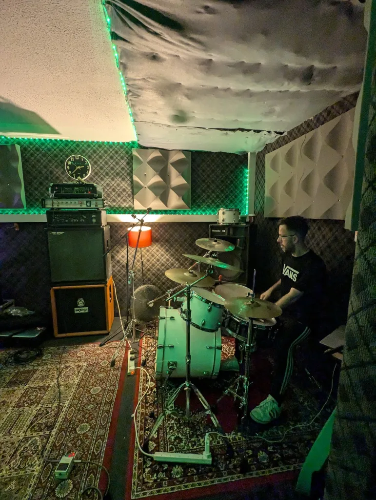 A man plays a white drum kit in a rehearsal studio lit with green LEDs, surrounded by acoustic panels and stacked guitar amplifiers.