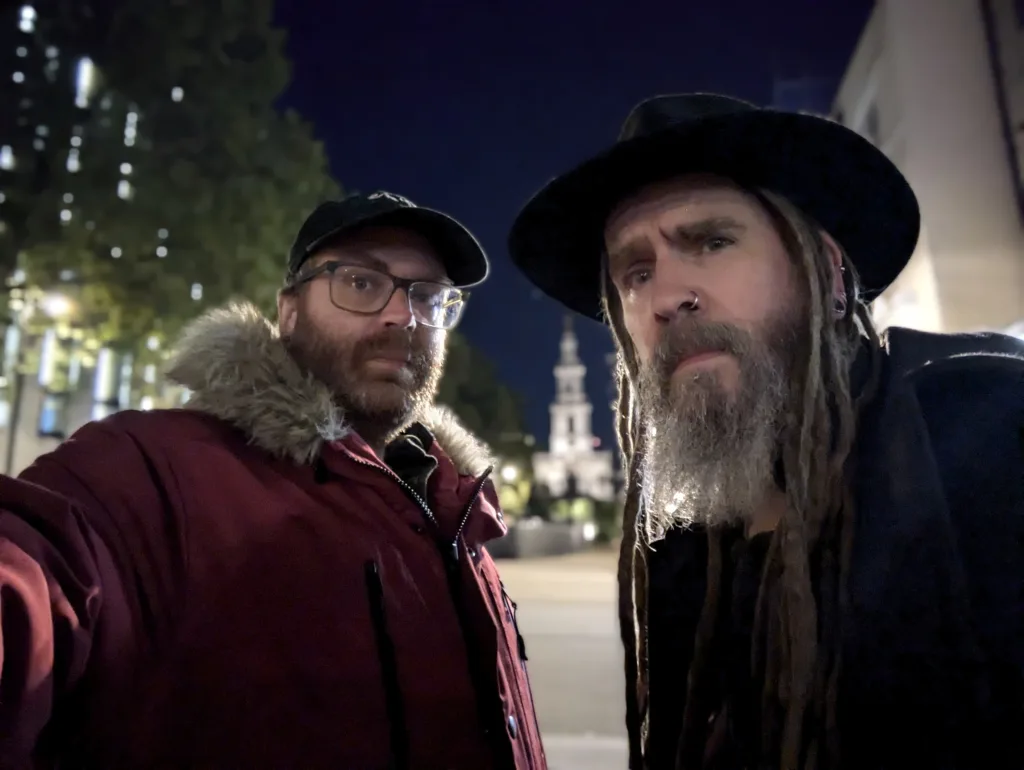 Two bearded men at night; one wearing a red parka and cap, the other a black wide-brimmed hat and long dreadlocks, with a lit tower behind them.