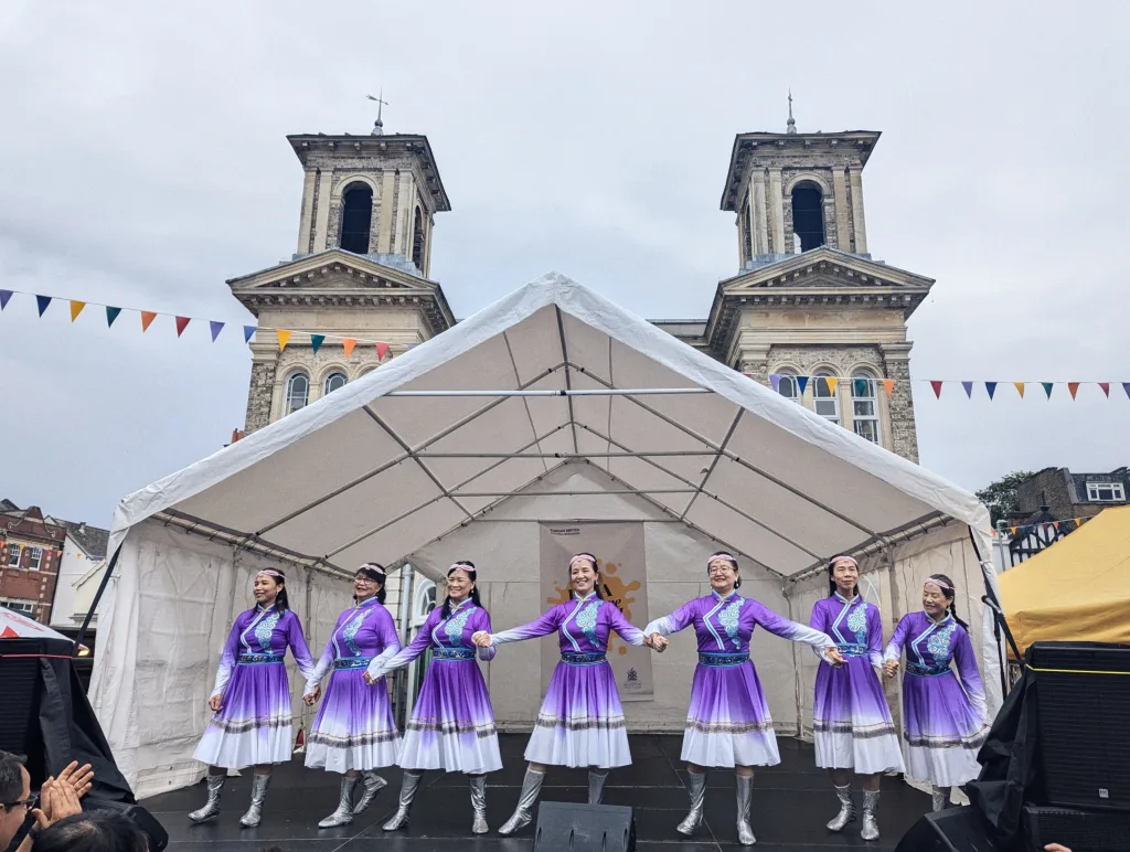 Seven women in purple and white traditional dresses dance hand-in-hand on a stage before a grand twin-towered stone building.