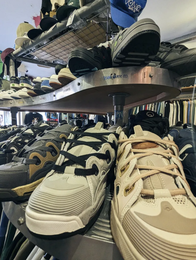 Close-up of chunky skate shoes on a metal display shelf in a skate shop, with baseball caps and clothing racks visible in the background.