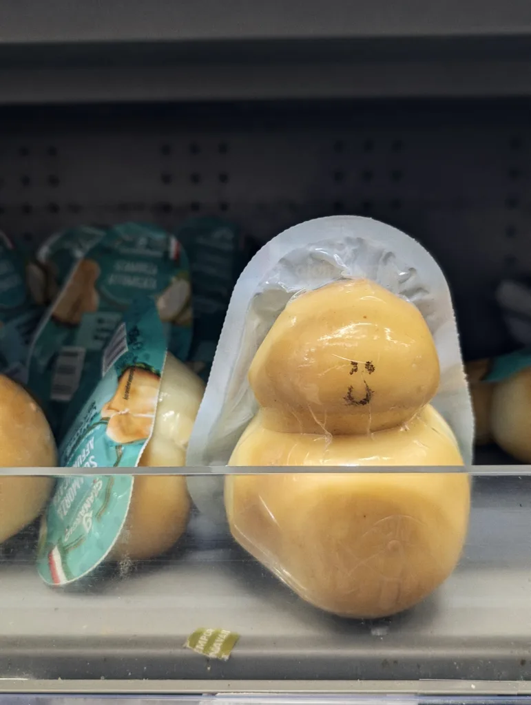 A vacuum-packed bulb of scamorza cheese on a supermarket shelf with small black marks that look like a tiny smiley face.