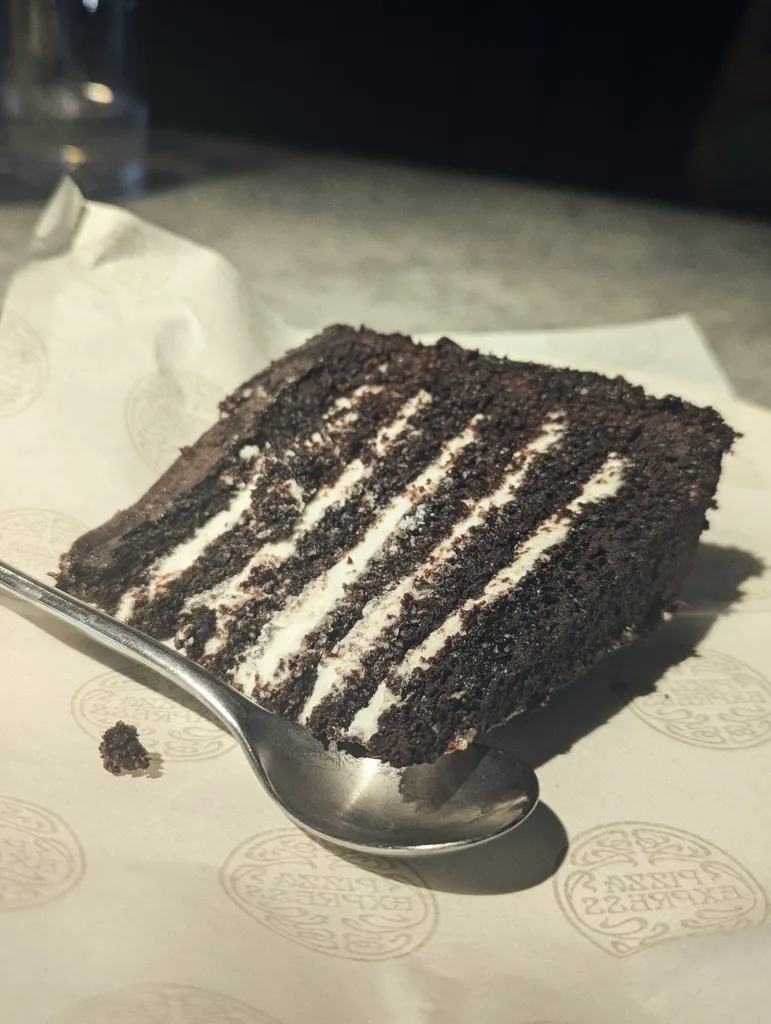 A slice of multi-layered chocolate cake with white cream filling on Pizza Express branded paper beside a silver spoon.