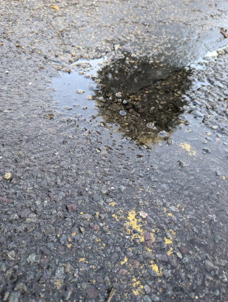 A small puddle of water on dark textured asphalt with yellow paint flecks.