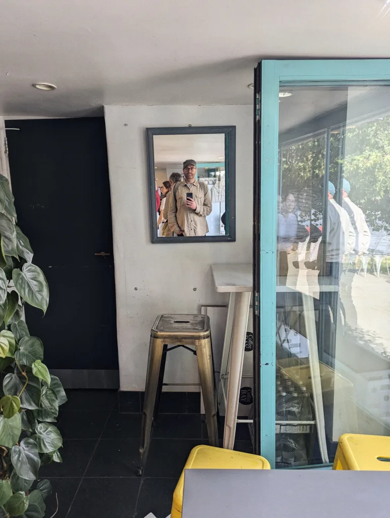 Mirror selfie of a man in a cap and beige shirt, reflected in a wall-mounted mirror above a metal stool in a cafe with a turquoise door frame.