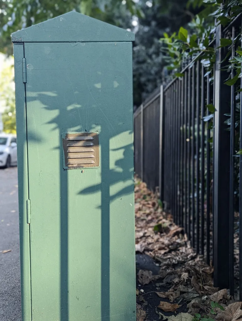 Green utility cabinet with a pointed top and small vent, cast in shadows next to a black metal fence and fallen brown leaves.