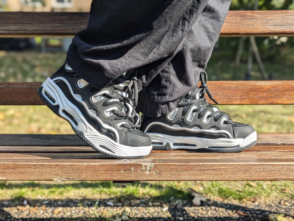Chunky black and white skate shoes with oversized laces and thick soles, worn on a wooden park bench.