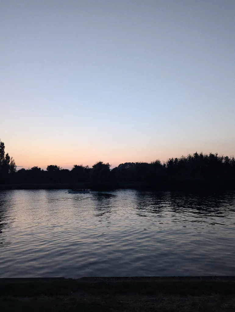 A small boat on a wide river at twilight, with trees silhouetted against a gradient sky of soft orange and blue.