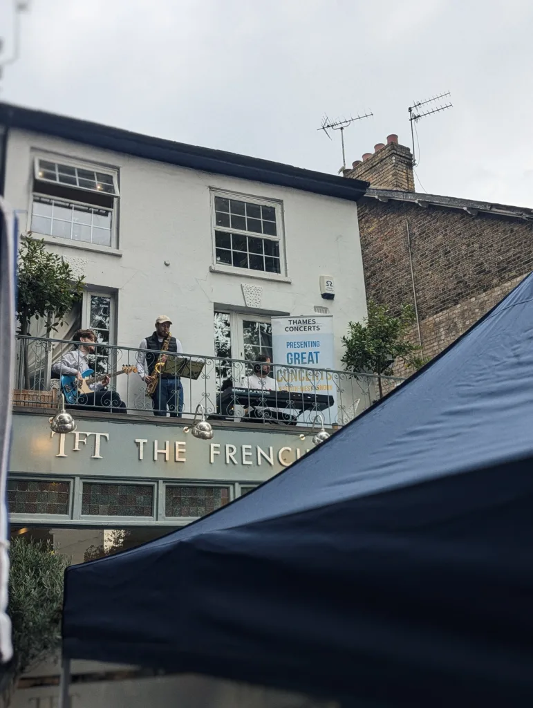 A jazz band with a saxophonist, bassist, and keyboardist performing on the balcony of The French pub under a Thames Concerts banner.