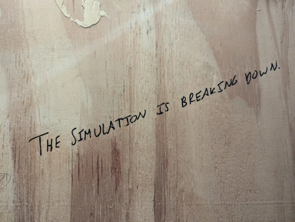 Handwritten black text on a textured wooden surface reads THE SIMULATION IS BREAKING DOWN.