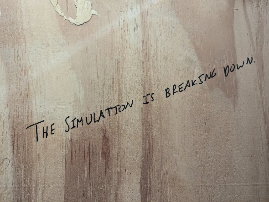 Handwritten message on a weathered wooden surface reading "THE SIMULATION IS BREAKING DOWN." in black marker.