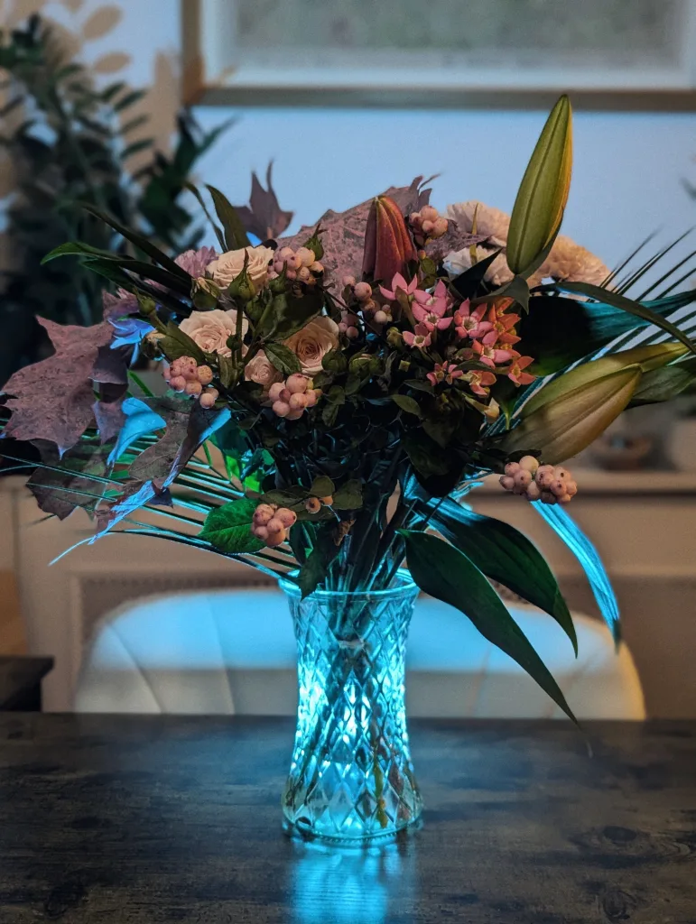 A bouquet of pink roses, berries, and lilies in a crystal vase illuminated by a cyan glow on a dark wooden table.