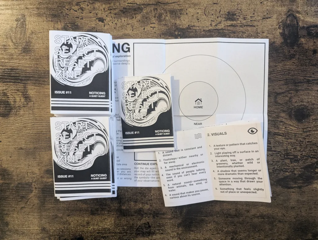 Stacks of Issue 11 of Start Select Reset Zine and an unfolded mapping prompt sheet on a textured wooden table.