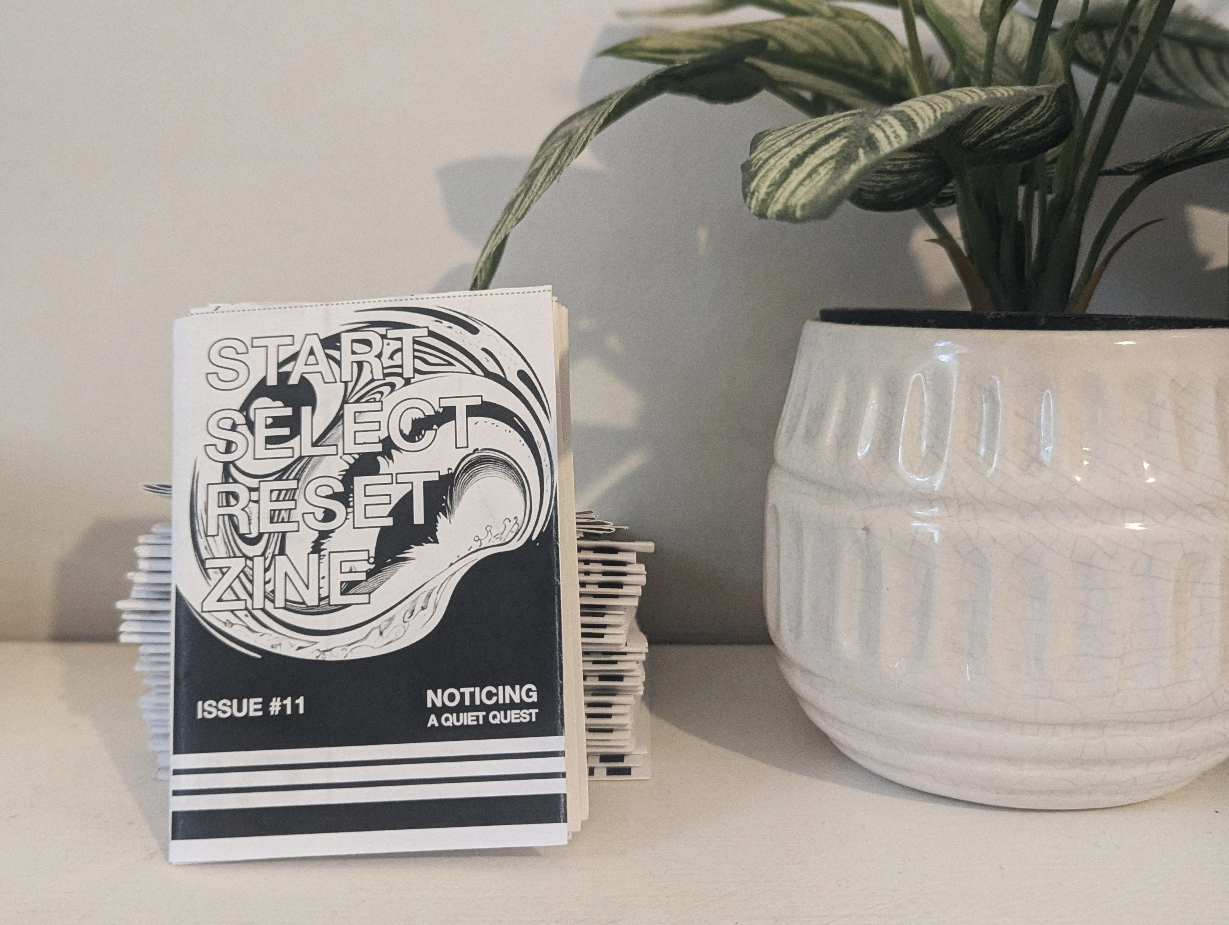 A stack of zines titled Start Select Reset Issue 11: Noticing a Quiet Quest, next to a leafy green plant in a white ceramic pot.