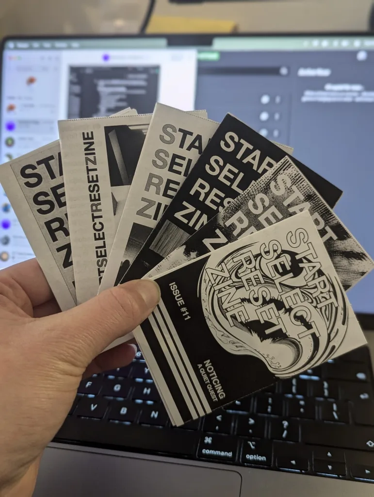 A hand holds a fanned stack of monochrome 'Start Select Reset' zines, including Issue 11, in front of an open laptop.