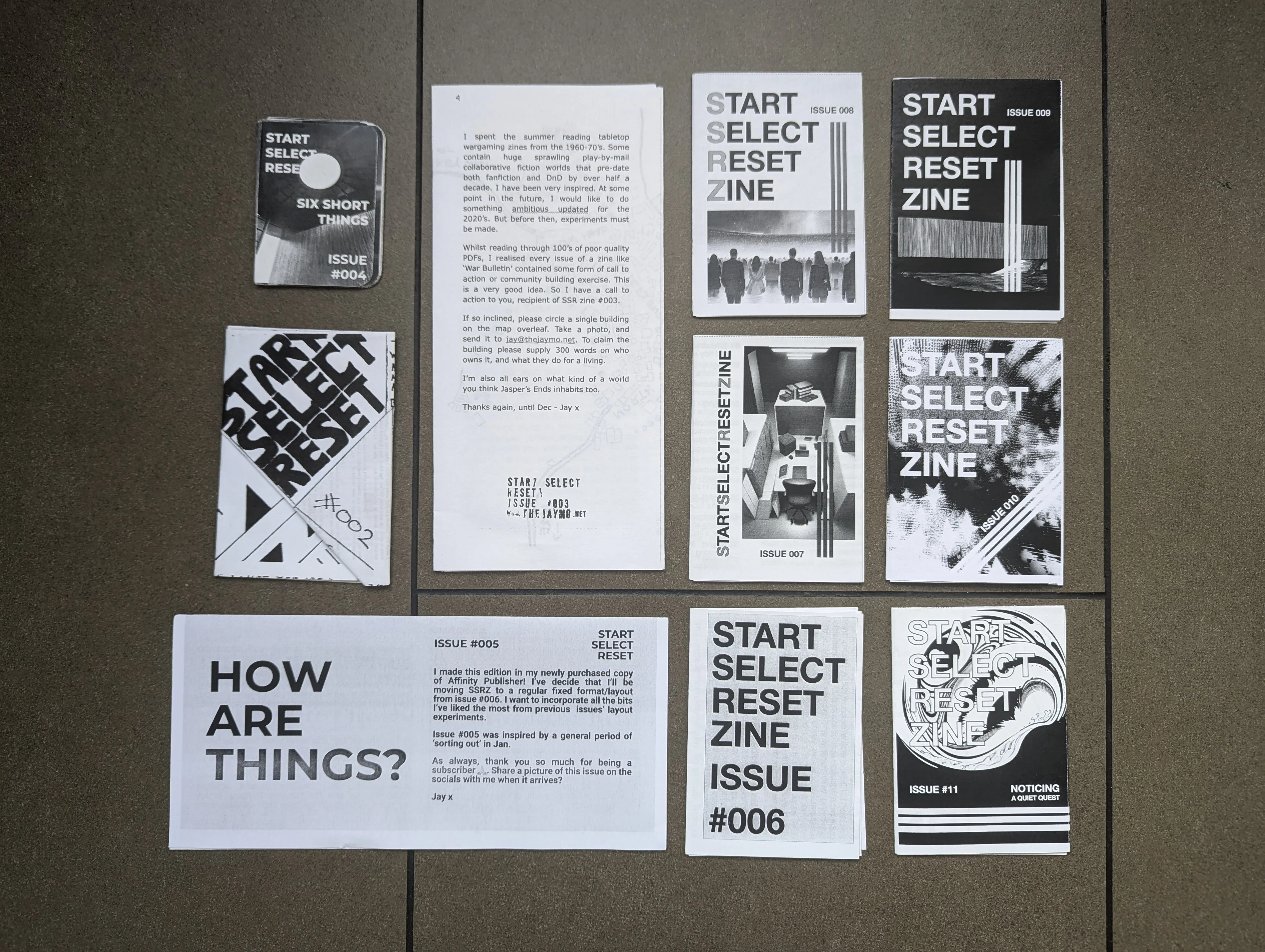 Various issues of the black and white START SELECT RESET zine laid out in a grid on a brown tiled floor.