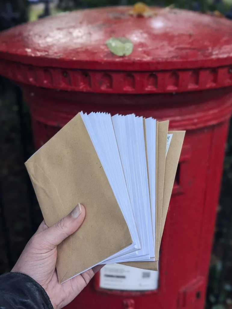 A hand fanning out a stack of white and brown envelopes in front of a red British pillar box.