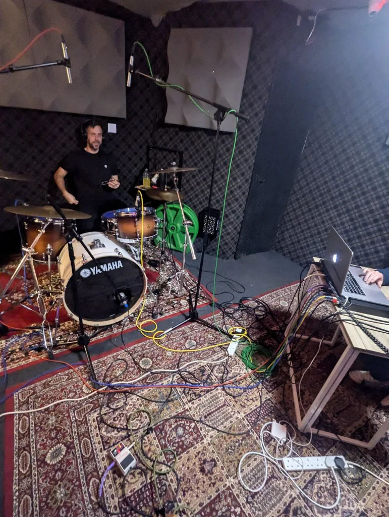 A man seated at a Yamaha drum kit in a music studio, surrounded by overhead microphones and a web of cables on patterned rugs.