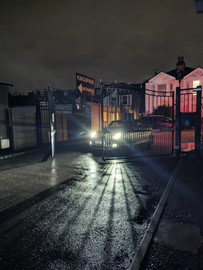 Headlights of a car behind a black metal gate reflecting on wet asphalt at night, beside a sign for Kingspark Entrance.