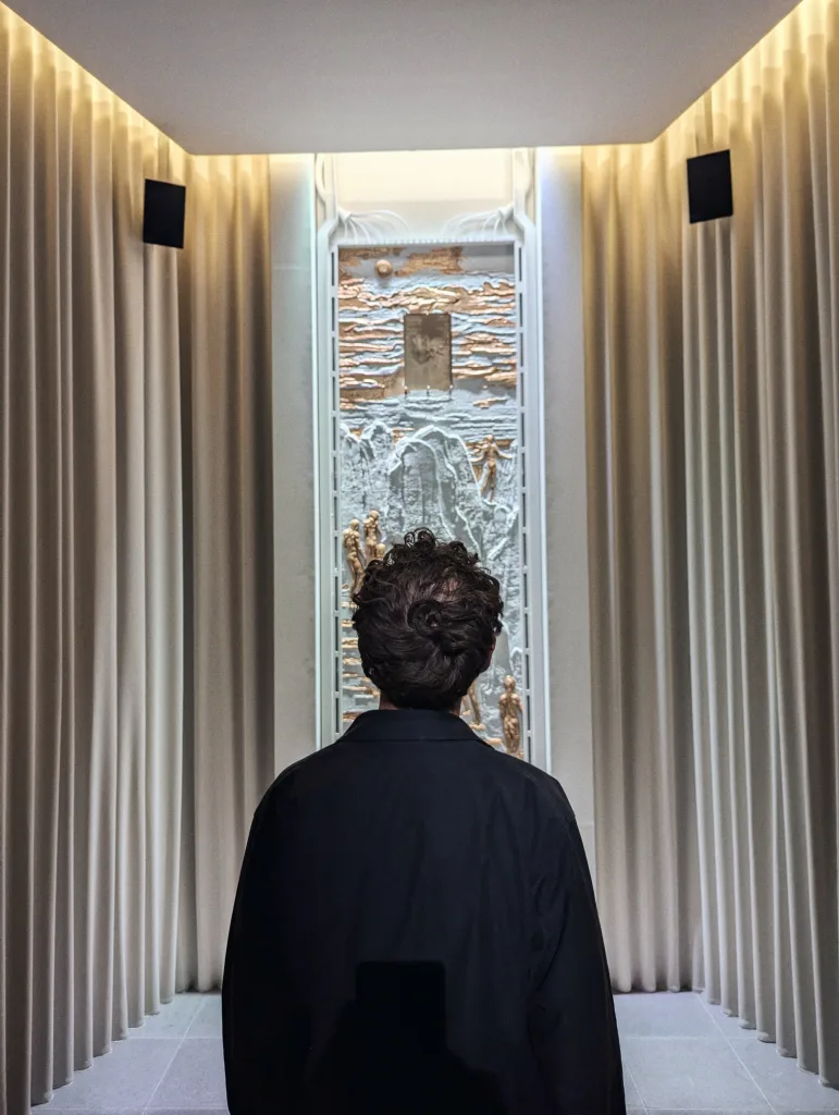 A person from behind looking at a vertical relief sculpture of human figures, framed by pleated beige curtains in a softly lit, narrow room.