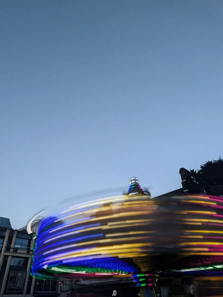 Colorful light trails from a spinning fairground ride in motion at dusk, set against urban buildings and a clear blue sky.