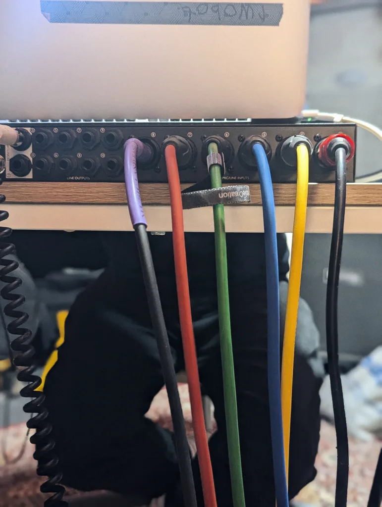 Purple, red, green, blue, and yellow audio cables plugged into the rear panel of a black interface on a desk.