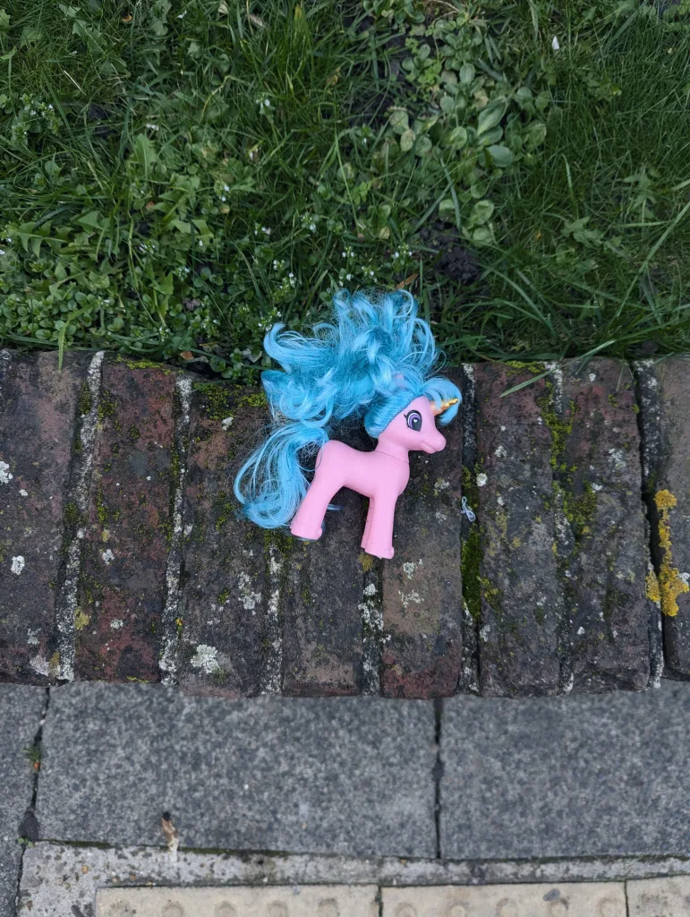 A pink unicorn toy with a tangled bright blue mane and tail lying on a mossy brick ledge between a lawn and a pavement.
