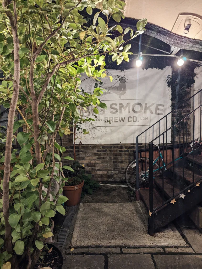 Big Smoke Brew Co. banner on a brick wall in a leafy courtyard at night with warm string lights, a bicycle, and stairs.