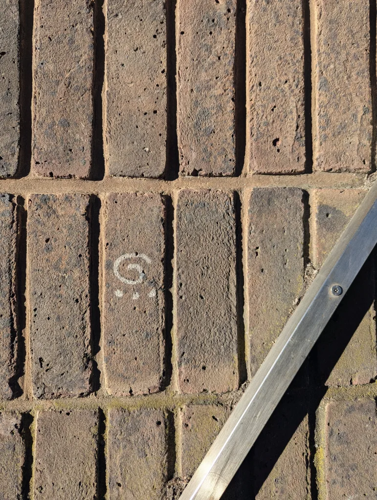 A small white chalk spiral and three dots drawn on a weathered brown brick wall, with a diagonal metal rail in the lower right.