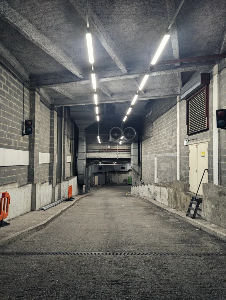 A concrete service ramp descending into an industrial tunnel, lit by parallel lines of overhead fluorescent lights against grey brick walls.